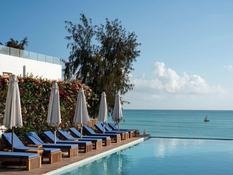 Piscina con sedie sdraio blu e ombrelloni accanto al mare sotto un cielo limpido.