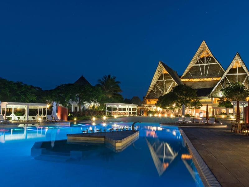 Piscine d'hôtel éclairée la nuit avec des bâtiments à toit triangulaire en arrière-plan