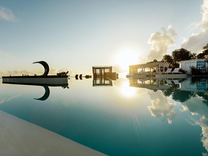 Piscine avec eau calme reflétant sculptures, palmiers et bâtiments au coucher du soleil.