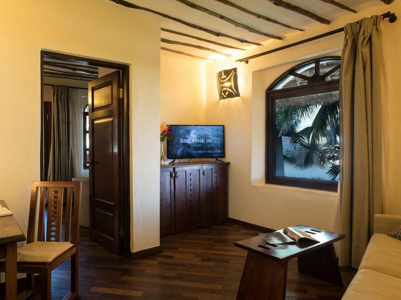 Coin salon d'hôtel confortable avec télévision, meubles en bois et vue sur des palmiers par la fenêtre.