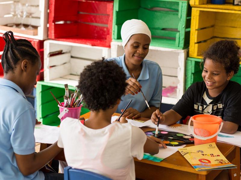 Des enfants et un adulte peignent ensemble à une table avec des caisses en bois colorées en arrière-plan.