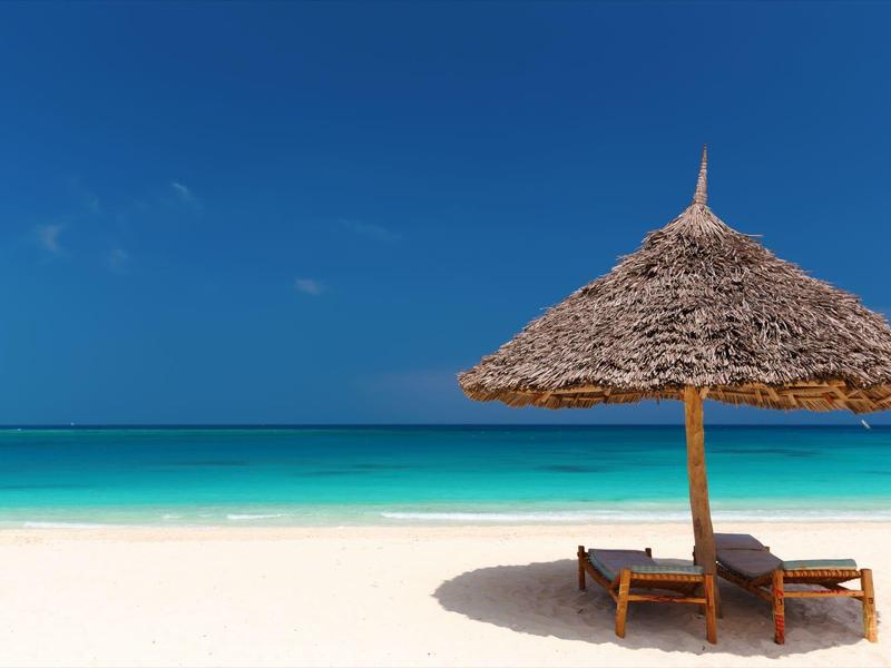 Strand mit weißem Sand, türkisfarbenem Meer und blauer Himmel, zwei Liegestühle unter Strohmantel.