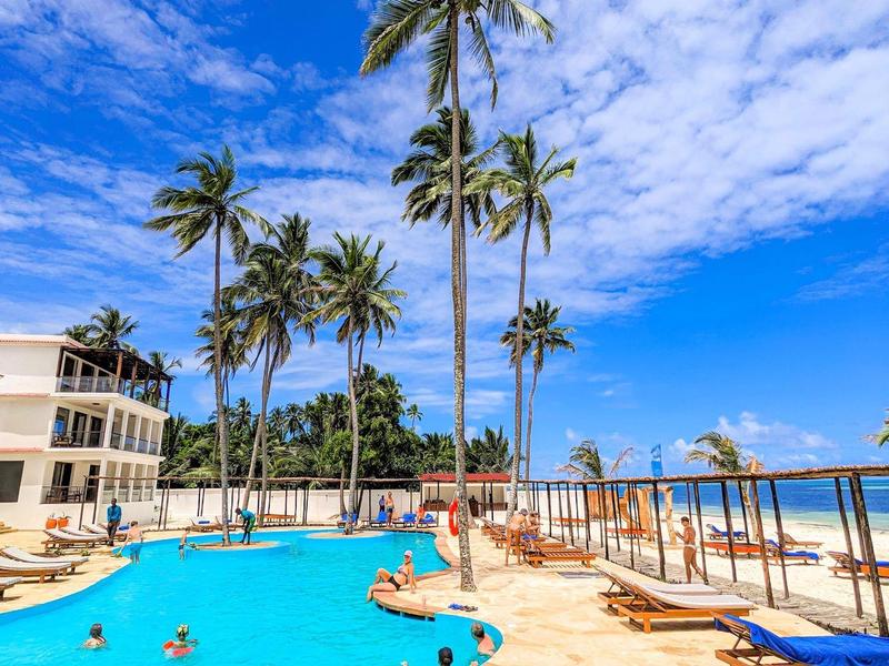 Schwimmbad mit Palmen, blauer Himmel und Liegestühlen auf beigem Steinboden am sonnigen Tag.