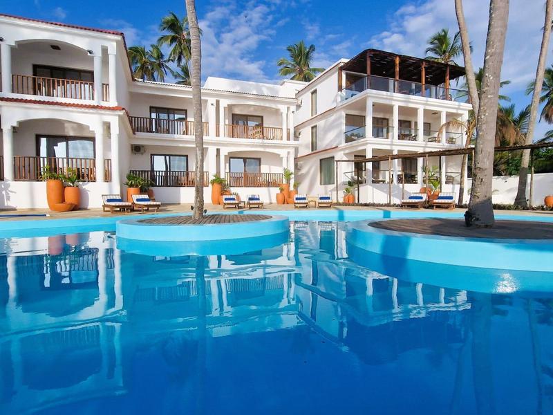 Großes weißes Resort mit mehreren Stockwerken, blauer Pool im Vordergrund, Palmen und klarer Himmel.