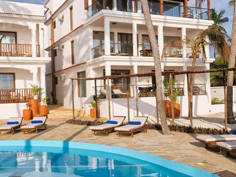 Mehrstöckiges Gebäude neben blauem Pool mit Sonnenliegen und Palmen unter blauem Himmel.