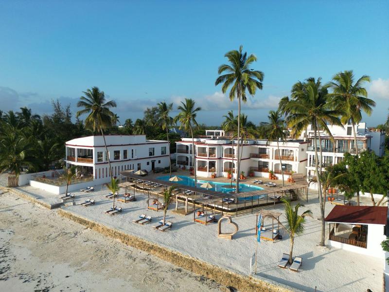Strandhotel mit Pool, Palmen und Sonnenschirmen an hellem Sandstrand unter klarem Himmel.