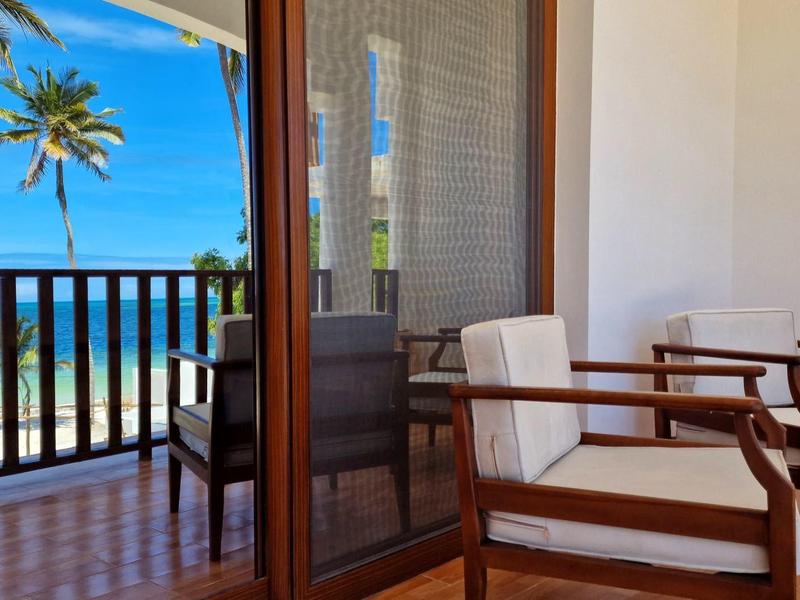 Balkon mit Holzgeländer, zwei Stühlen und Blick auf Palmen und Meer bei blauem Himmel.