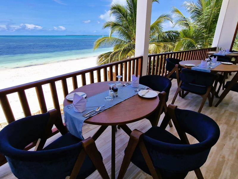 Terrassenbereich mit Holzboden, runden Tischen, blauen Stühlen und Blick auf Palmen und Meer.