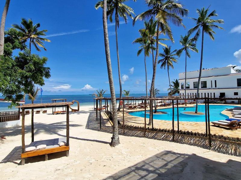 Strand mit Sand, Liegen, hohen Palmen und Pool vor weißem Gebäude unter blauem Himmel.