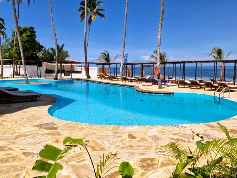 Ein blauer Pool mit Palmen und Pflanzen umgeben, Meer und Himmel im Hintergrund.