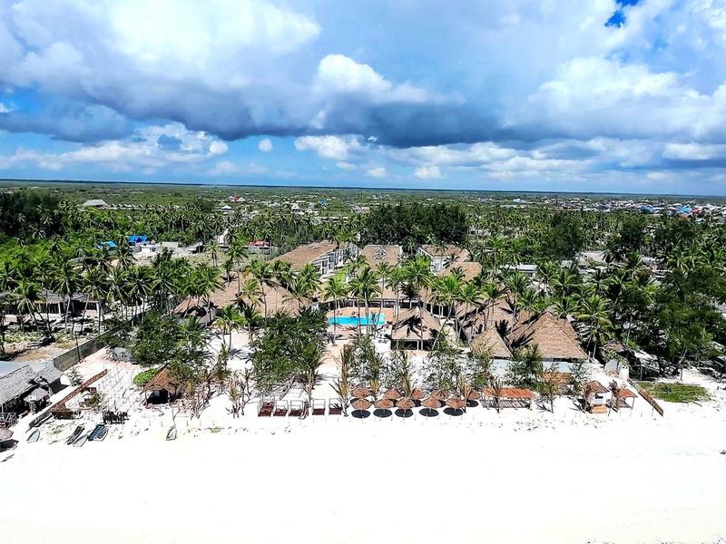 Blick auf tropischen Strand mit weißen Sand, Palmen und mehreren Strohdachbungalows unter blauem Himmel.