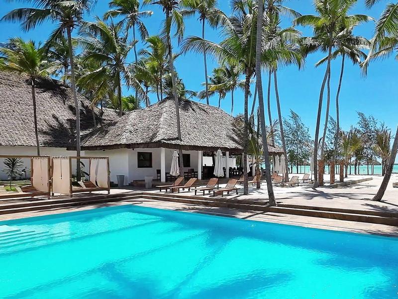 Blauer Pool vor weißen Bungalows mit Strohdächern, umgeben von hohen Palmen und blauem Himmel.