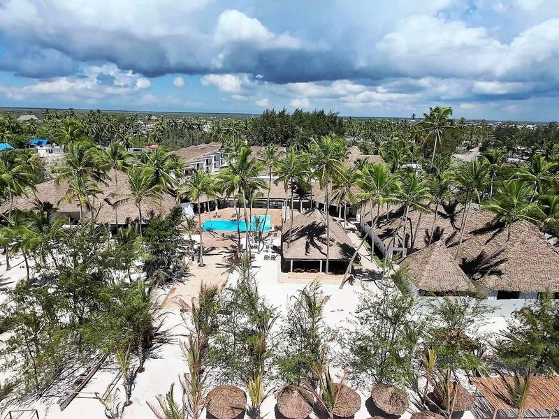 Übersichtliches Resort mit Strohdächern, Palmen und blauem Pool vor bewölktem Himmel am Strand.