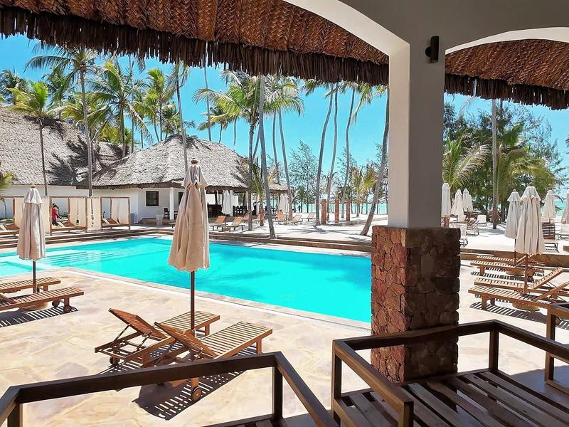Poolbereich mit Liegestühlen, Sonnenschirmen und Palmen unter blauem Himmel.