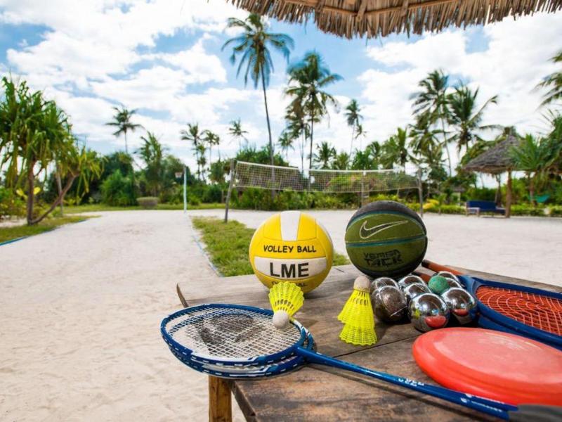 Sandplatz mit Tennis- und Beachvolleyball-Ausrüstung, Palmen und Blauem Himmel im Hintergrund.