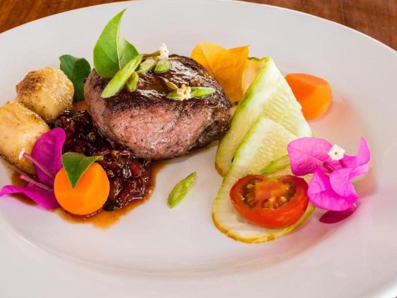 Einzeln angerichtetes Gericht mit gebratenem Fleisch, Gemüse, süßer Soße und essbaren Blüten auf weißem Teller.