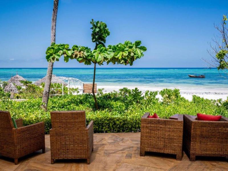 Vier Korbstühle mit roten Kissen stehen auf Terrasse mit Blick auf weißen Sandstrand und blaues Meer.