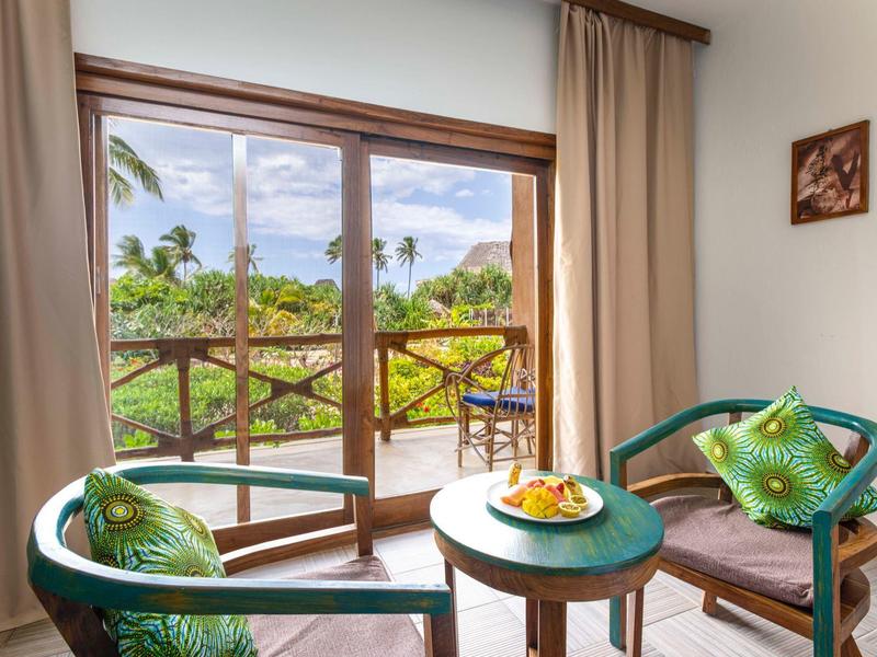 Helles Zimmer mit Holzfenstern, zwei Stühlen, Tisch und Blick auf grüne Pflanzen und blauen Himmel.