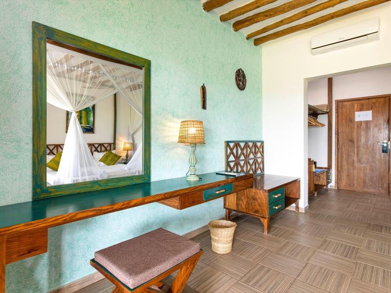 Hellgrünes Zimmer mit Spiegel, Holzbank, Hocker, Lampe und braunem Türbereich im Hintergrund.