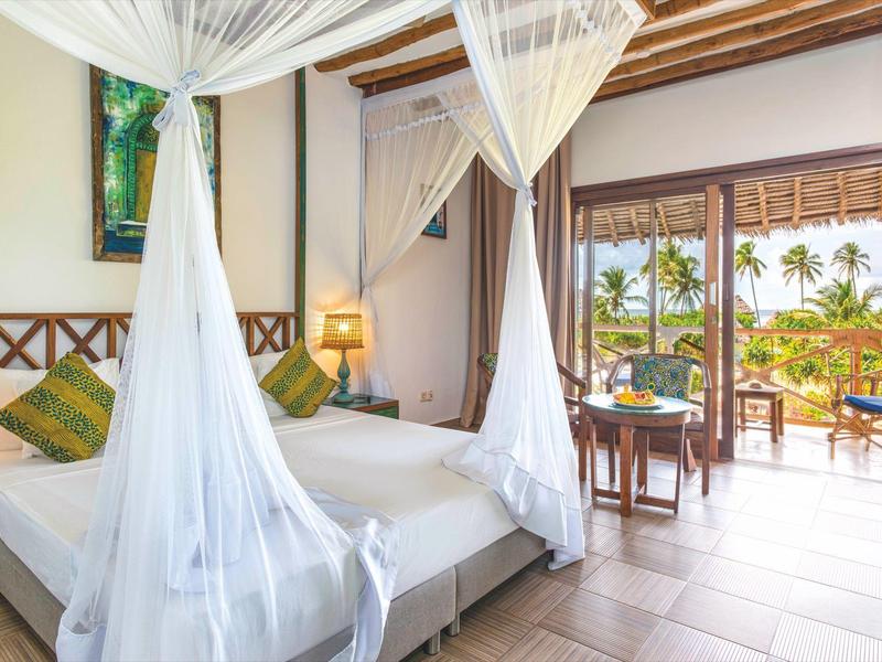 Helles Schlafzimmer mit Himmelbett, Holzbalken, Blick auf Terrasse und Palmenlandschaft draußen.