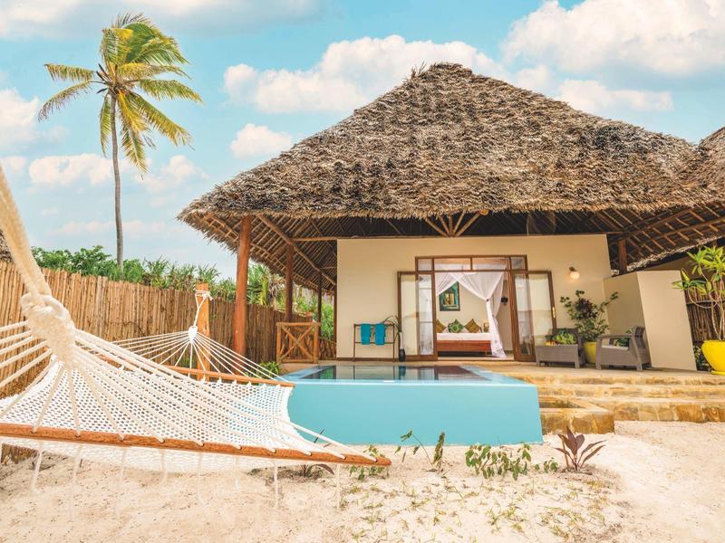 Strandhaus mit Strohdach, Pool, Hängematte und Palmen bei sonnigem Himmel am weißen Sandstrand.