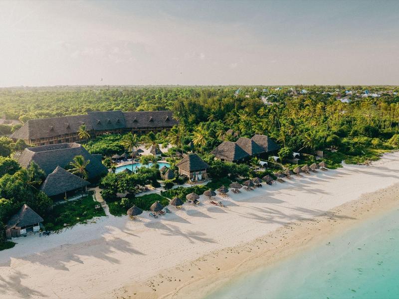 Strand mit weißem Sand, türkisfarbenem Wasser und mehreren kleinen Gebäuden im grünen Hinterland.