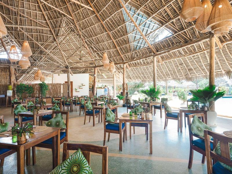 Offenes Restaurant mit Holzstühlen, grünen Kissen und hohem, strohgedecktem Dach.