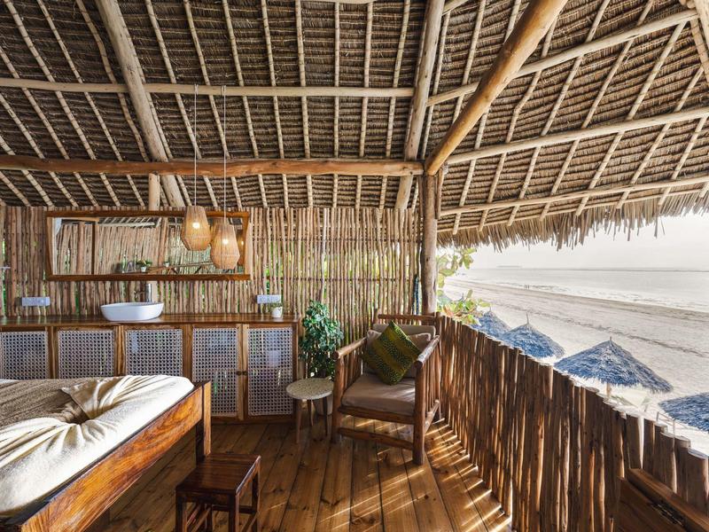 Offene Holz- und Strohhütte mit Bett, Stühlen, hängender Lampe und Blick aufs Meer.