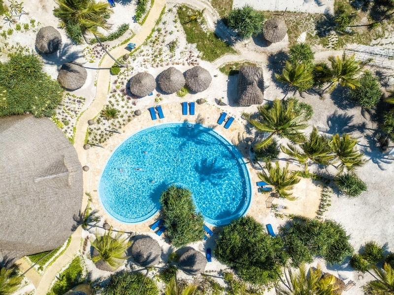 Herzförmiger Pool mit blauen Liegestühlen, Palmen und Strohdächern in tropischer Gartenanlage.