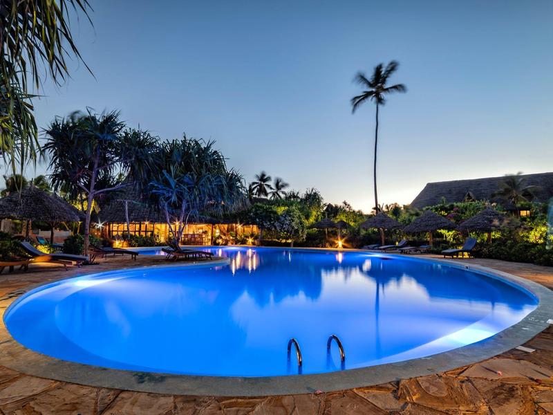 Runder beleuchteter Pool bei Dämmerung, umgeben von Palmen und Garten mit Sitzbereich.