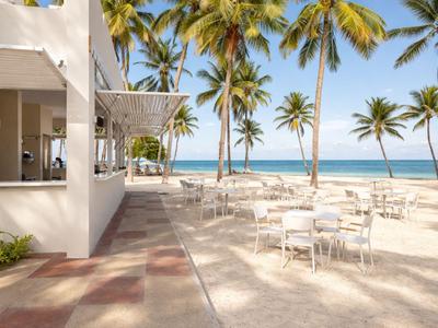 Prázdná venkovní část restaurace na pláži s bílými stoly a palmami v pozadí.
