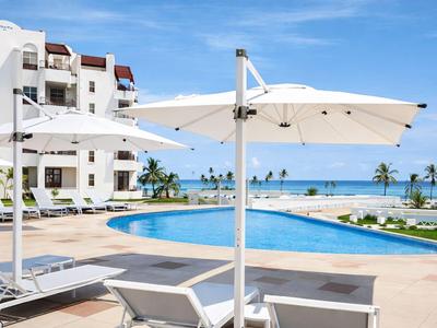 Basen hotelowy z leżakami i białymi parasolami, morze i palmy w tle.