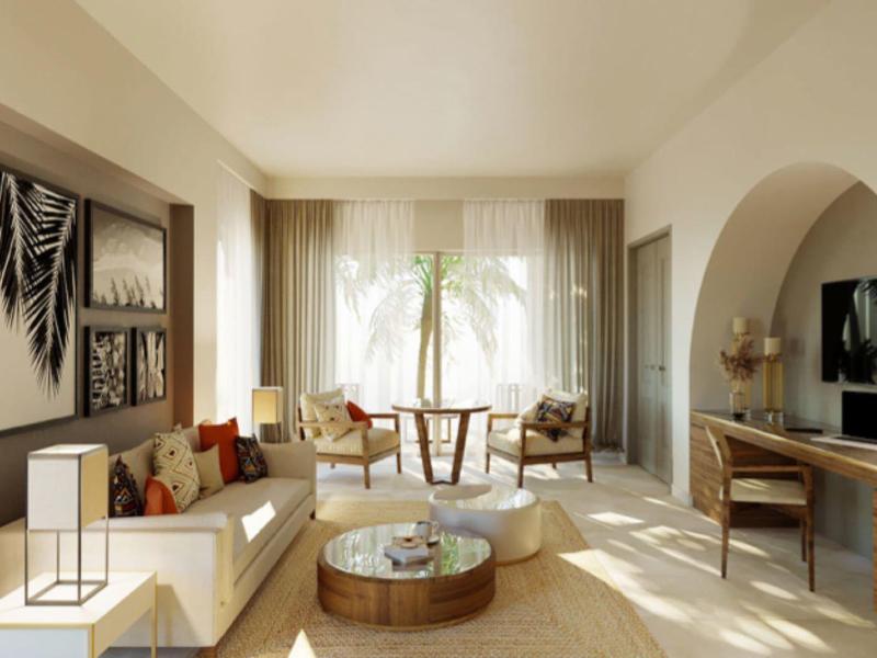 Jasny salon z beżowymi ścianami, narożną sofą, okrągłym stolikiem kawowym i oknem z zasłonami.