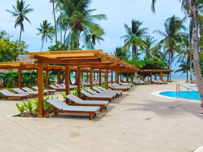 Strand mit Liegen unter Holzpergolen und Blick auf den Pool und Palmen.