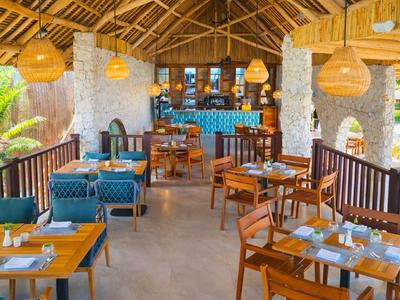 Offenes Restaurant mit Holzmöbeln, blauen Sitzpolstern und Bambuslampen unter einem Strohdach.