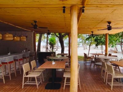 Freiluftrestaurant mit Holzdach, Tischen und Stühlen, Barbereich und Blick auf Strand.