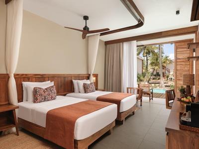 Camera d'hotel con due letti, pannelli in legno scuro, ventilatore a soffitto e vista sulla piscina.