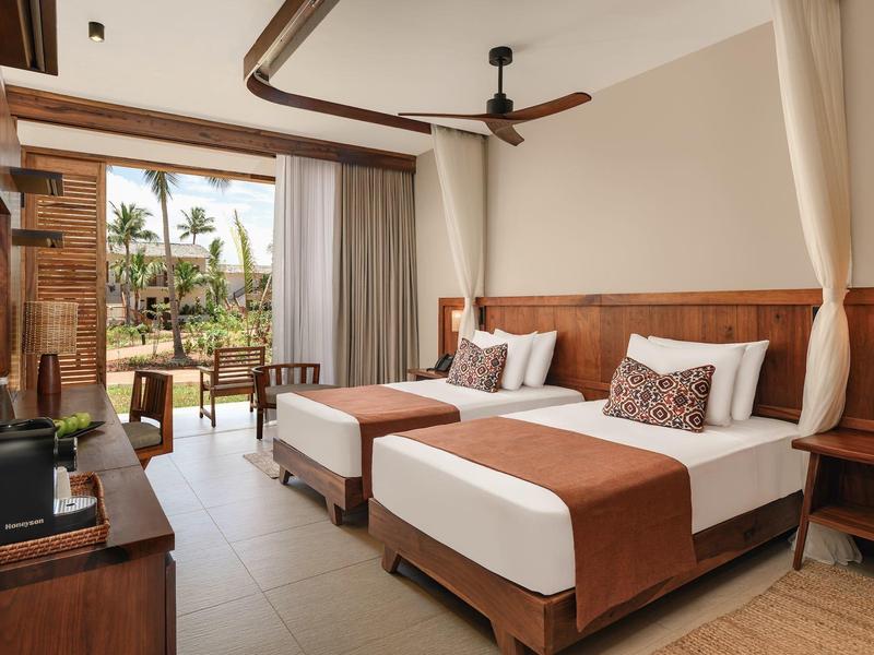 Camera d'hotel con due letti singoli, mobili in legno e accesso al balcone con vista sulle palme.