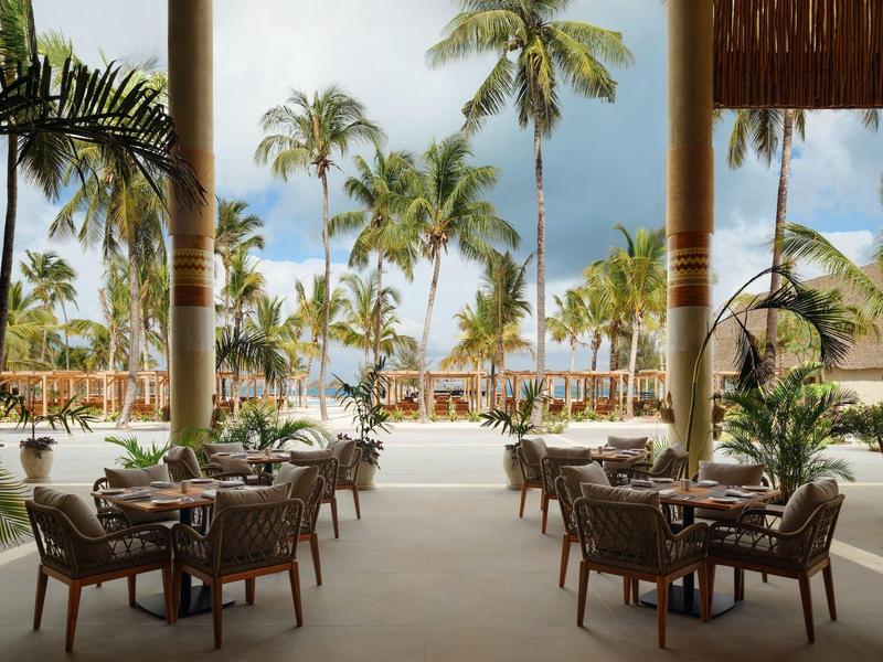 Vista da area esterna coperta con sedie e tavoli verso palme tropicali e edifici.