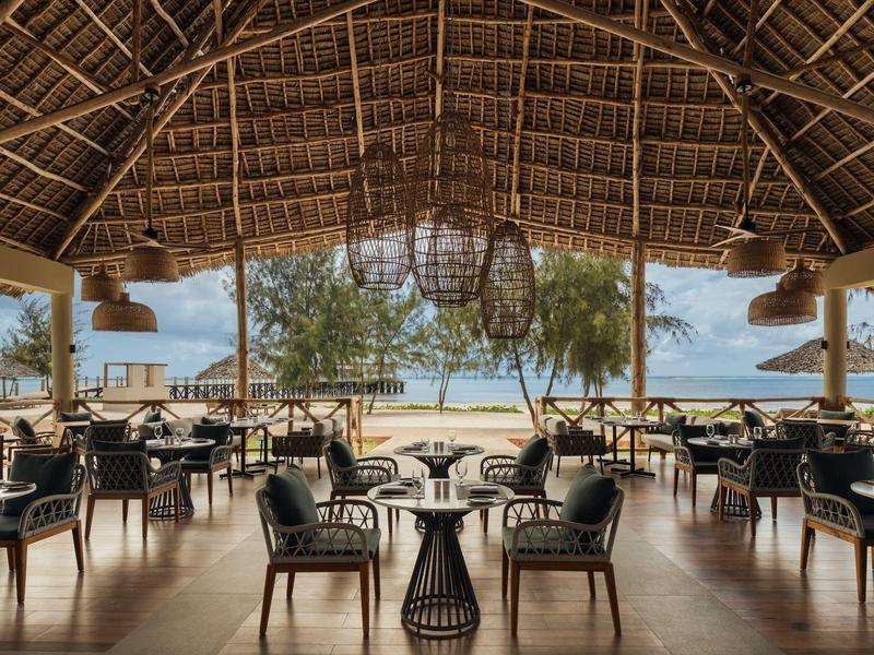 Bar sulla spiaggia aperta con sedie e tavoli sotto un tetto di paglia con vista sul mare.