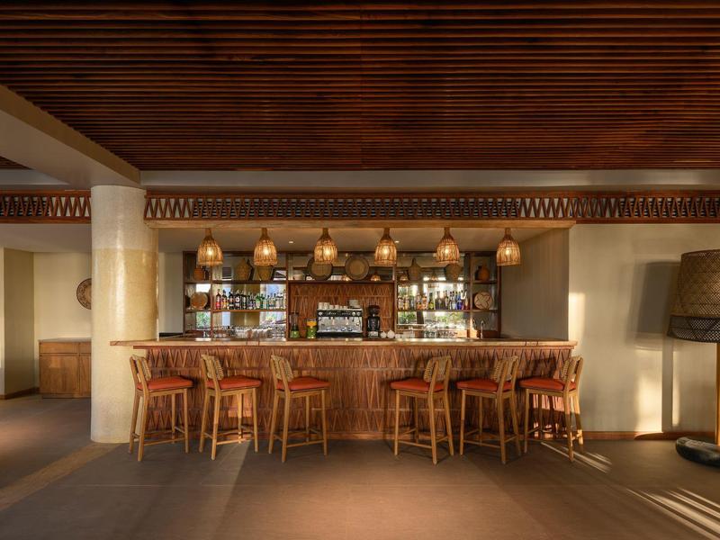Bar dell'hotel con bancone in legno, sedie in vimini e illuminazione calda, circondato da un interno moderno.