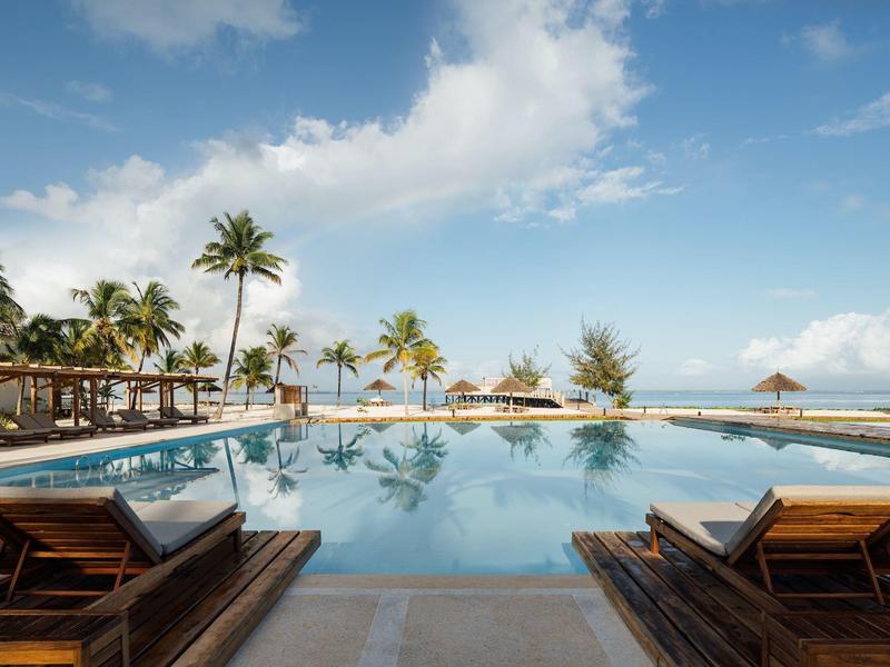 Vista di una piscina con lettini, palme e mare sullo sfondo con cielo limpido.