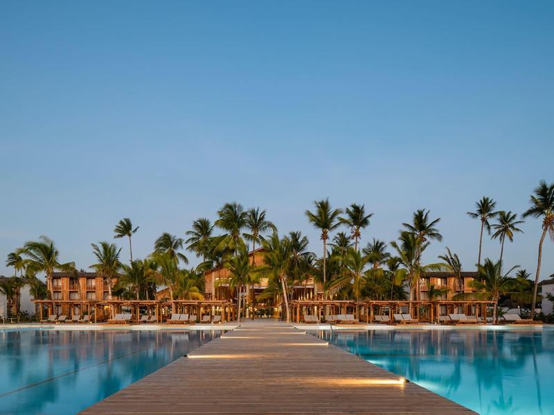 Molo conduce a un hotel con piscina e palme sotto un cielo azzurro limpido.