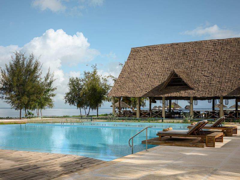 Area piscina con lettini e padiglione coperto vicino all'acqua sotto un cielo blu.
