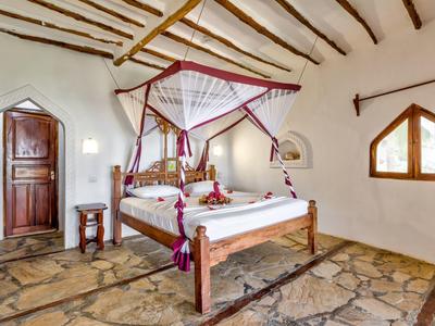 Helles Schlafzimmer mit Holzbalken, Steinfussboden, Himmelbett und roten Vorhängen am Fenster.