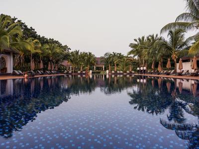 A long, calm pool with palm trees and lounge chairs on both sides under a clear sky.