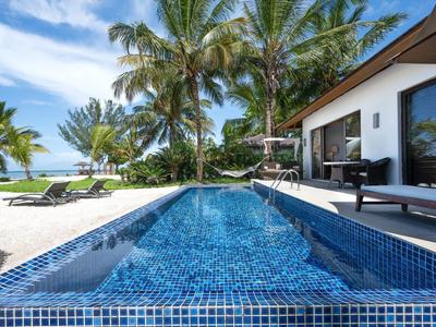 Moderner Pool mit blauen Kacheln, Palmen, Liegestühlen und Haus am weißen Sandstrand unter blauem Himmel.