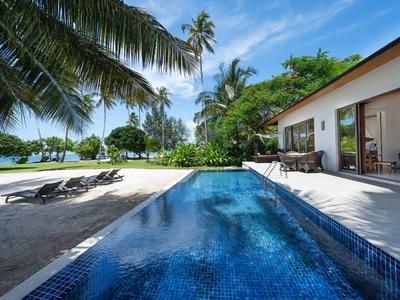 Moderner Pool mit blauen Fliesen neben Haus, Sandstrand, Palmen und blauem Himmel im Hintergrund.
