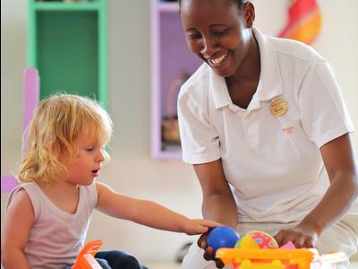 Ein kleines blondes Mädchen spielt mit buntem Spielzeugauto, neben lachender Betreuerin.