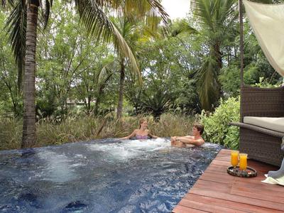 Ein Außenpool mit Holzdeck, Sofa, Handtuch und Blick auf grüne Bäume und Palmen.